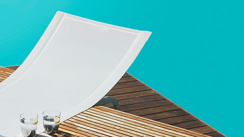 White sunlounger, water glasses and a book on a wooden deck with a swimming pool in the background